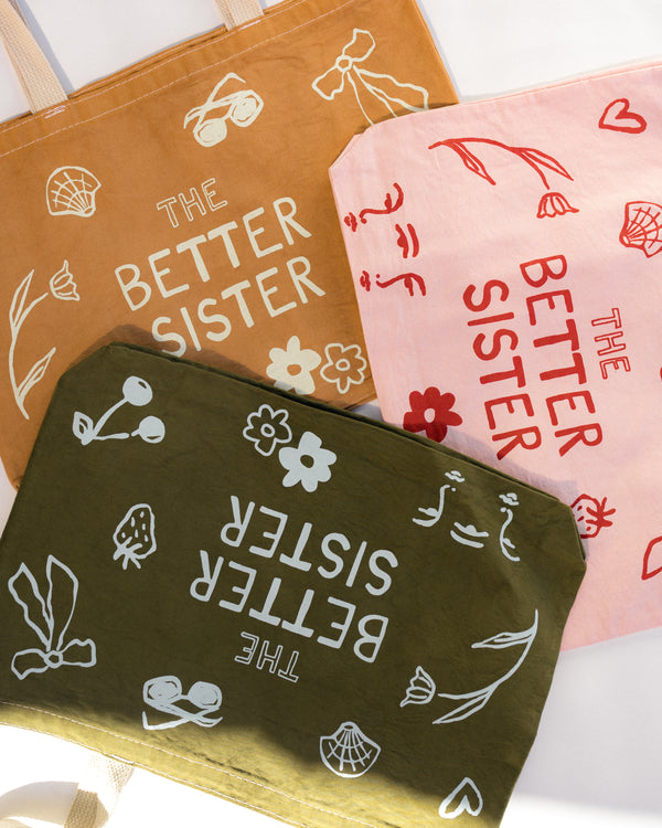 Three fabric tote bags arranged diagonally on a white background, each with a different base color: tan, pink, and olive green. Each bag features white or red hand-drawn doodles and the text 'THE BETTER SISTER' printed upside down.
